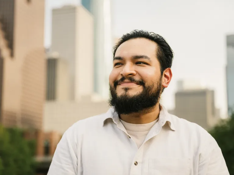 A confident Mexican man wearing a dark shirt and jacket, standing outdoors with a city skyline in the background. He gazes directly at the camera, exuding professionalism and self-assurance.