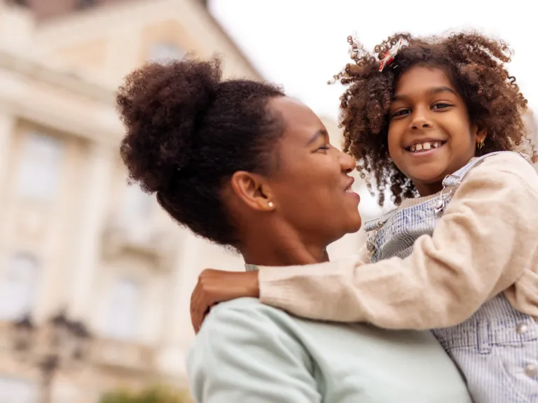 A mother and her young daughter share a joyful moment outdoors, with the mother lifting the daughter in her arms, both smiling and enjoying their time together.