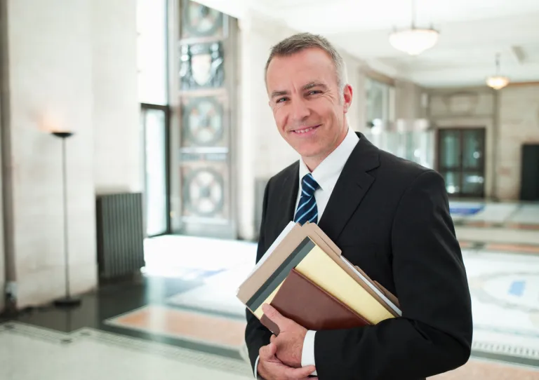 Middle-aged white male lawyer in a dark business suit and tie, standing in the bright, modern lobby of a law firm or courthouse, smiling confidently with arms loosely at his sides, and a briefcase or legal documents visible in the background.