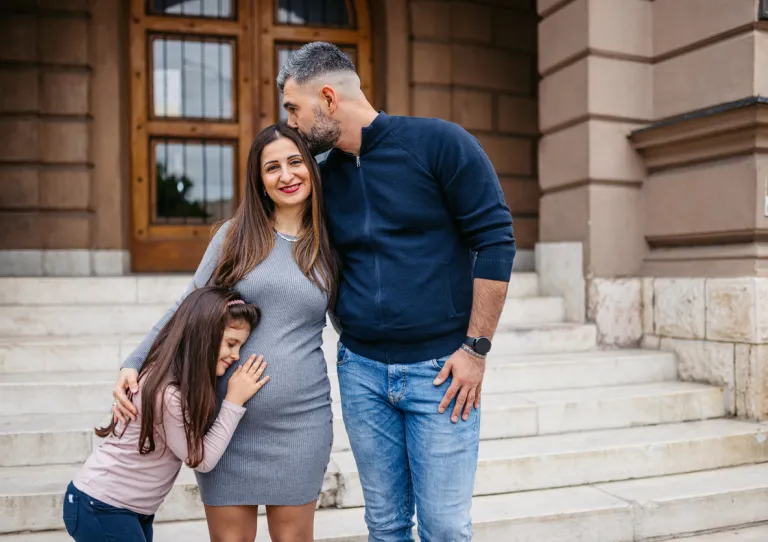 A father tenderly kisses his pregnant wife’s head while their young daughter embraces her mother’s belly, all sharing a joyful, intimate moment together.