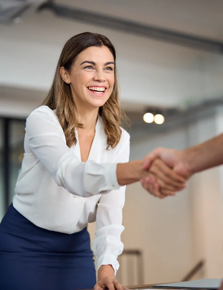 A happy, professional mid-aged businesswoman in a tailored blazer smiles warmly as she shakes hands with a client in a bright office setting, signaling a successful meeting or agreement.