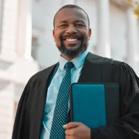 Mature Black male lawyer in a dark suit and tie, standing in a city-street setting with tall buildings behind him, holding a briefcase as he walks confidently toward a courthouse.