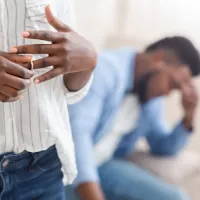 woman in foreground taking off wedding ring while man in background has head in hands